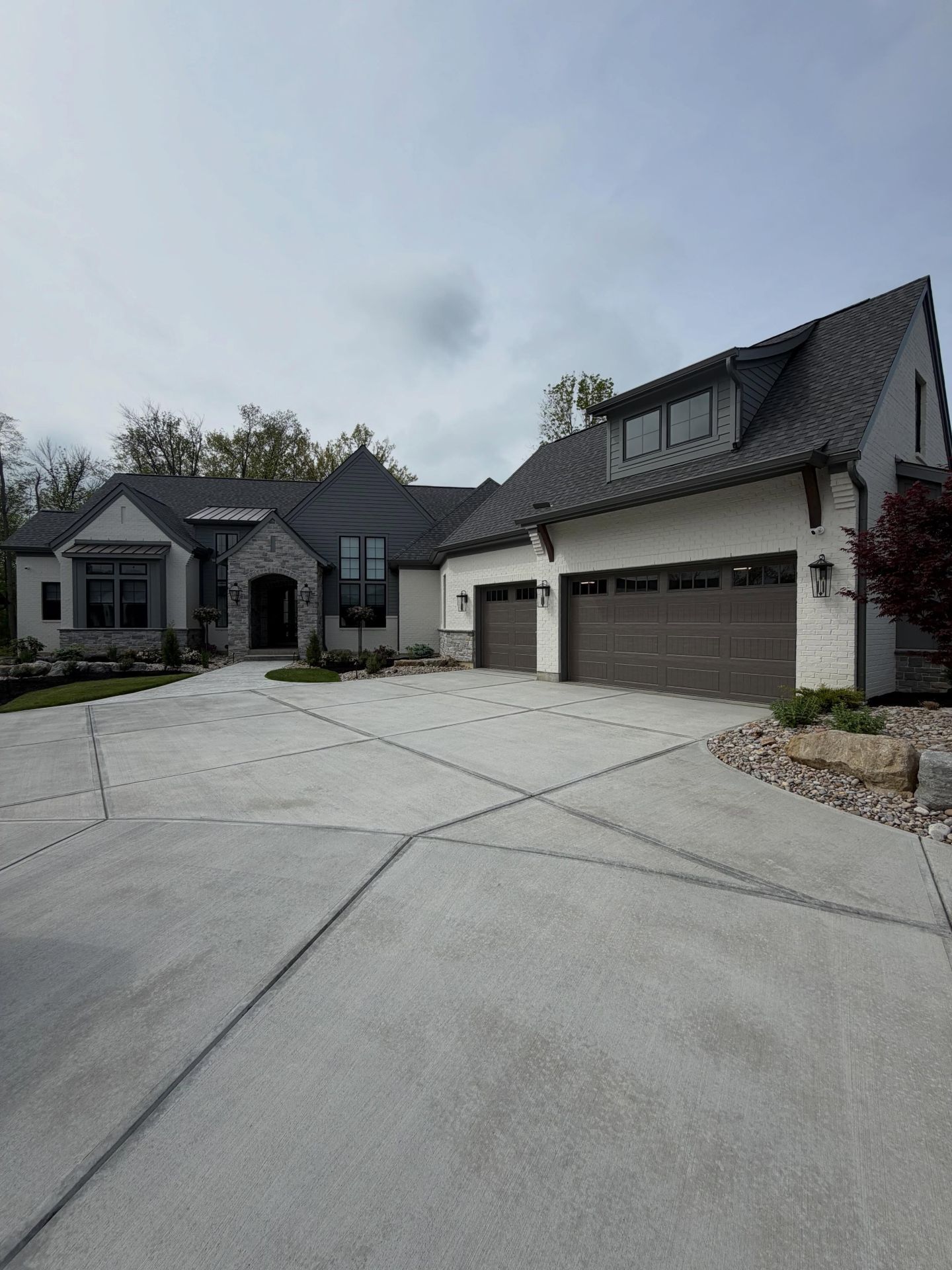 Large modern suburban house with gray roof, three-car garage, and wide concrete driveway under a cloudy sky