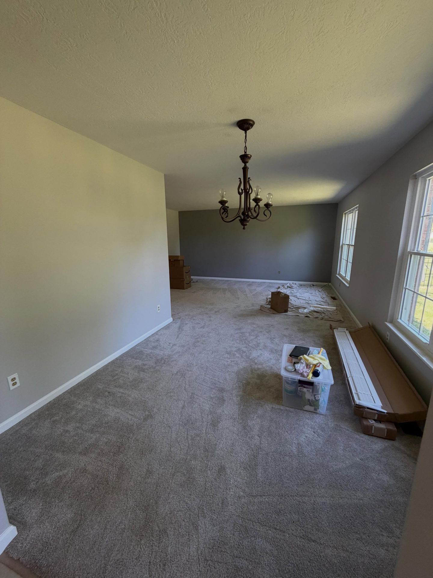 Empty carpeted dining room with chandelier, beige walls, and boxes near the window on the right.