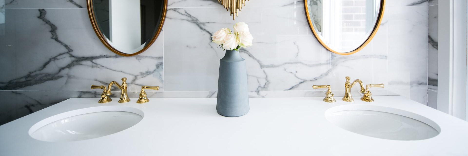 Salle de bains avec marbre blanc, miroirs ovales, robinetterie dorée et vase de fleurs blanches.