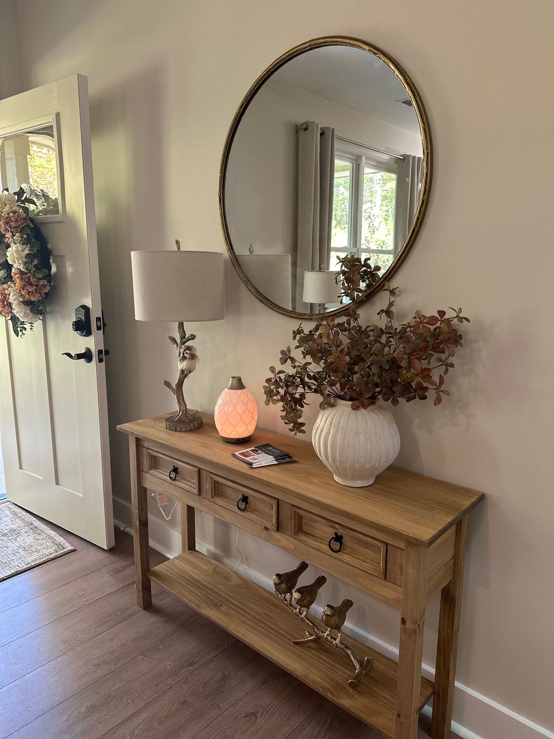 A hallway with a wooden table , mirror , lamp and vase of flowers.