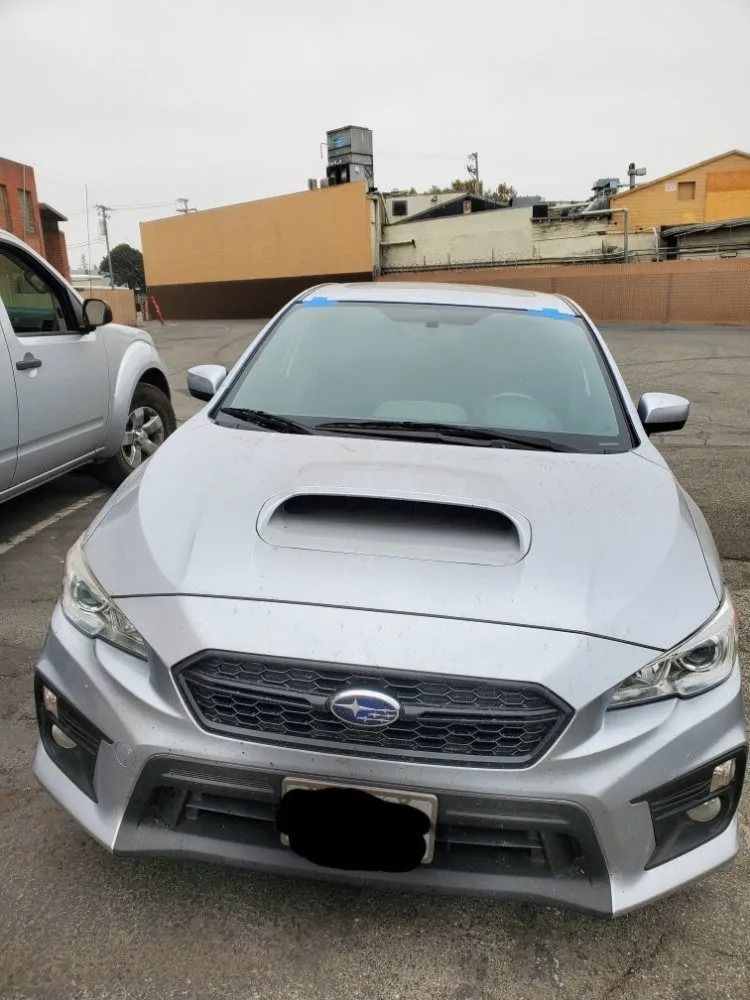 Silver Subaru WRX parked in a lot; features hood scoop, black grill, blue tape on windshield.