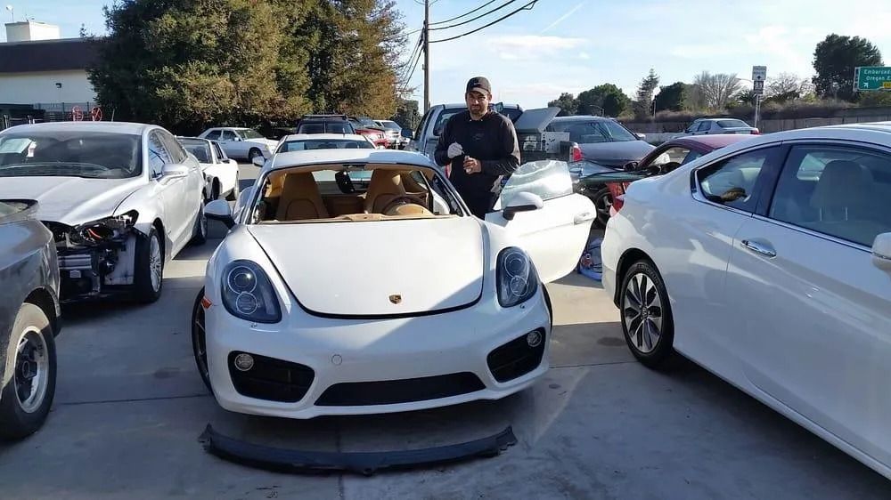 White Porsche Cayman surrounded by other cars in a parking lot; a person stands near the car.