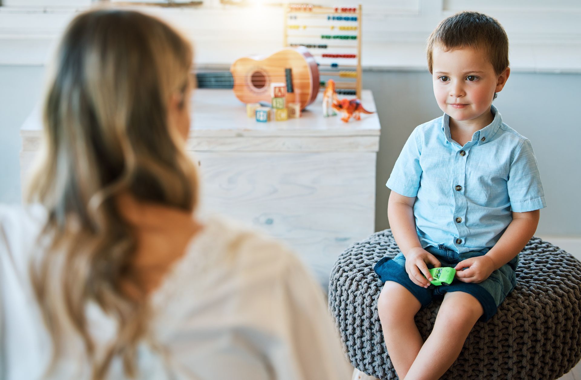 A young boy is sitting on an ottoman talking to a woman.