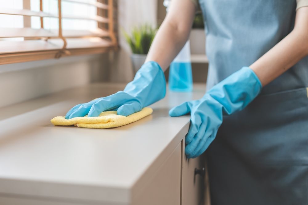 Person wearing blue gloves wiping a white countertop with a yellow cloth, cleaning.