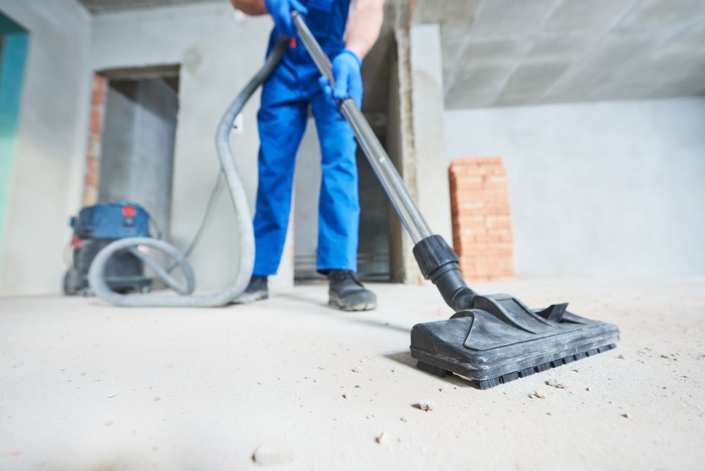 Construction worker in blue overalls vacuuming a dusty concrete floor with a large vacuum.