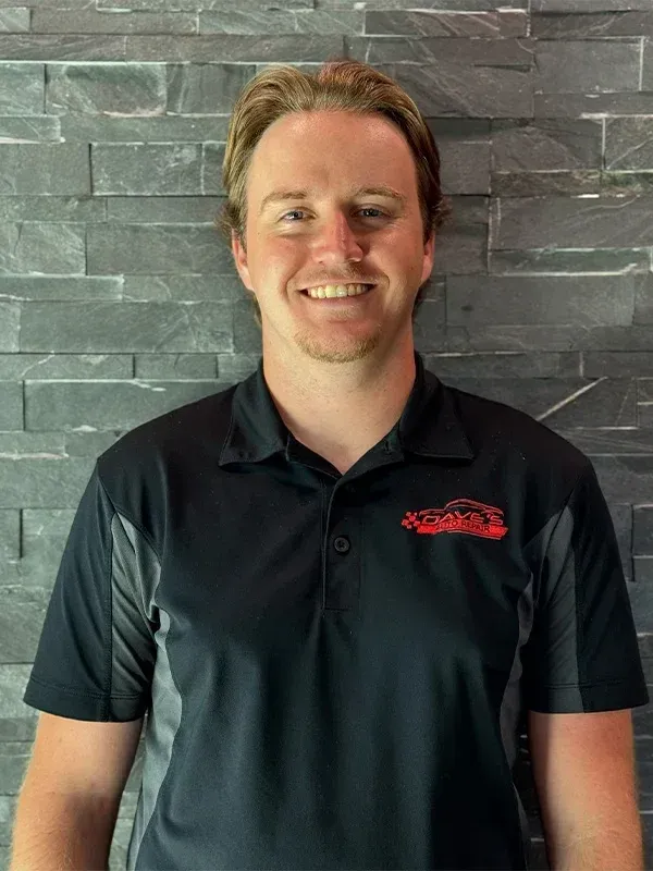 Man with blond hair smiling, wearing a black polo shirt with a logo, standing in front of a gray brick wall. | Dave's Auto Repair