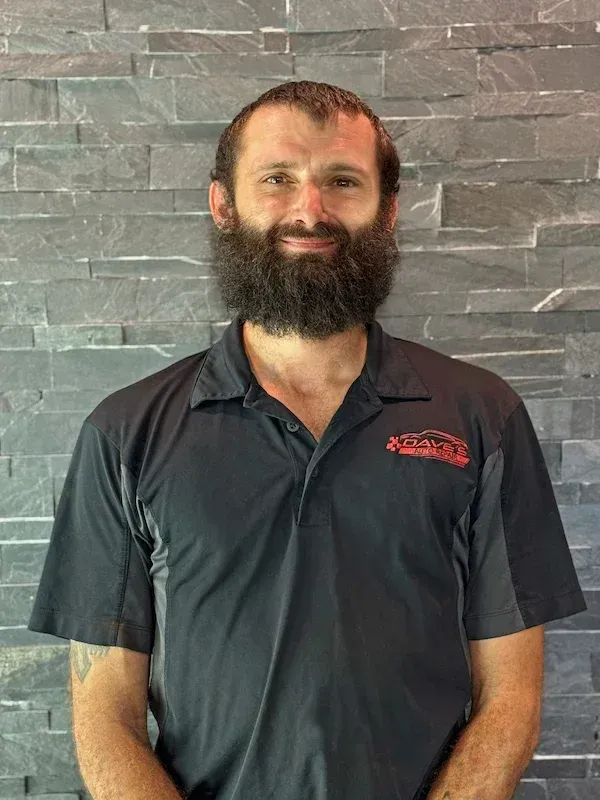Man with beard wearing black polo shirt smiles in front of a gray stone wall. | Dave's Auto Repair