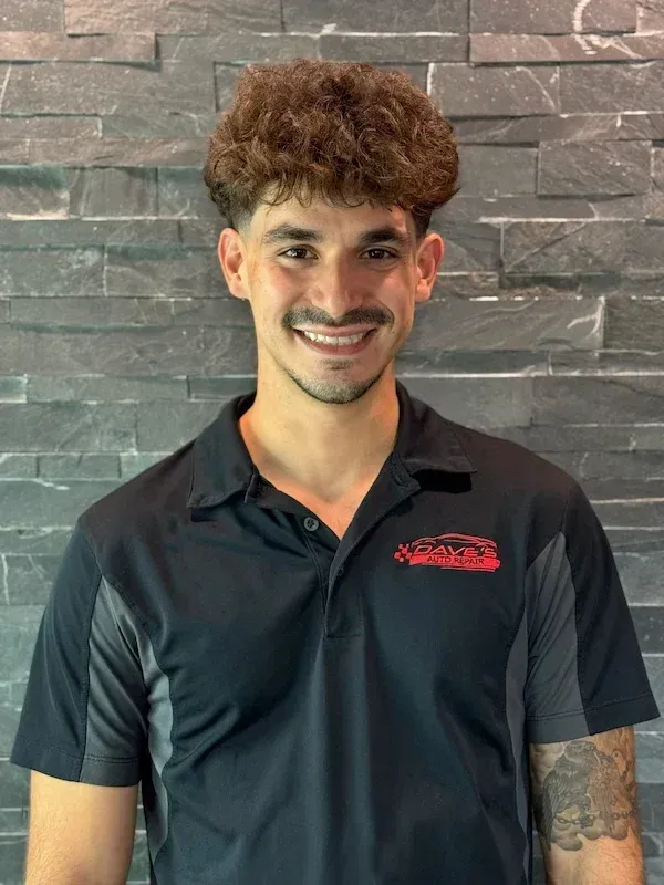 Man with curly hair and a smiling expression, wearing a black and gray polo shirt. Brick wall background. | Dave's Auto Repair