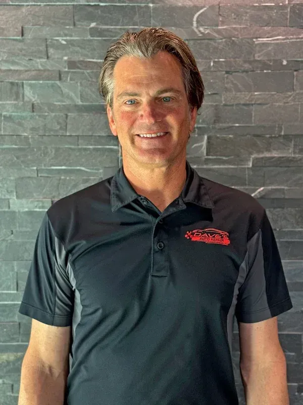 Man in black polo shirt smiling, standing in front of a gray brick wall. | Dave's Auto Repair