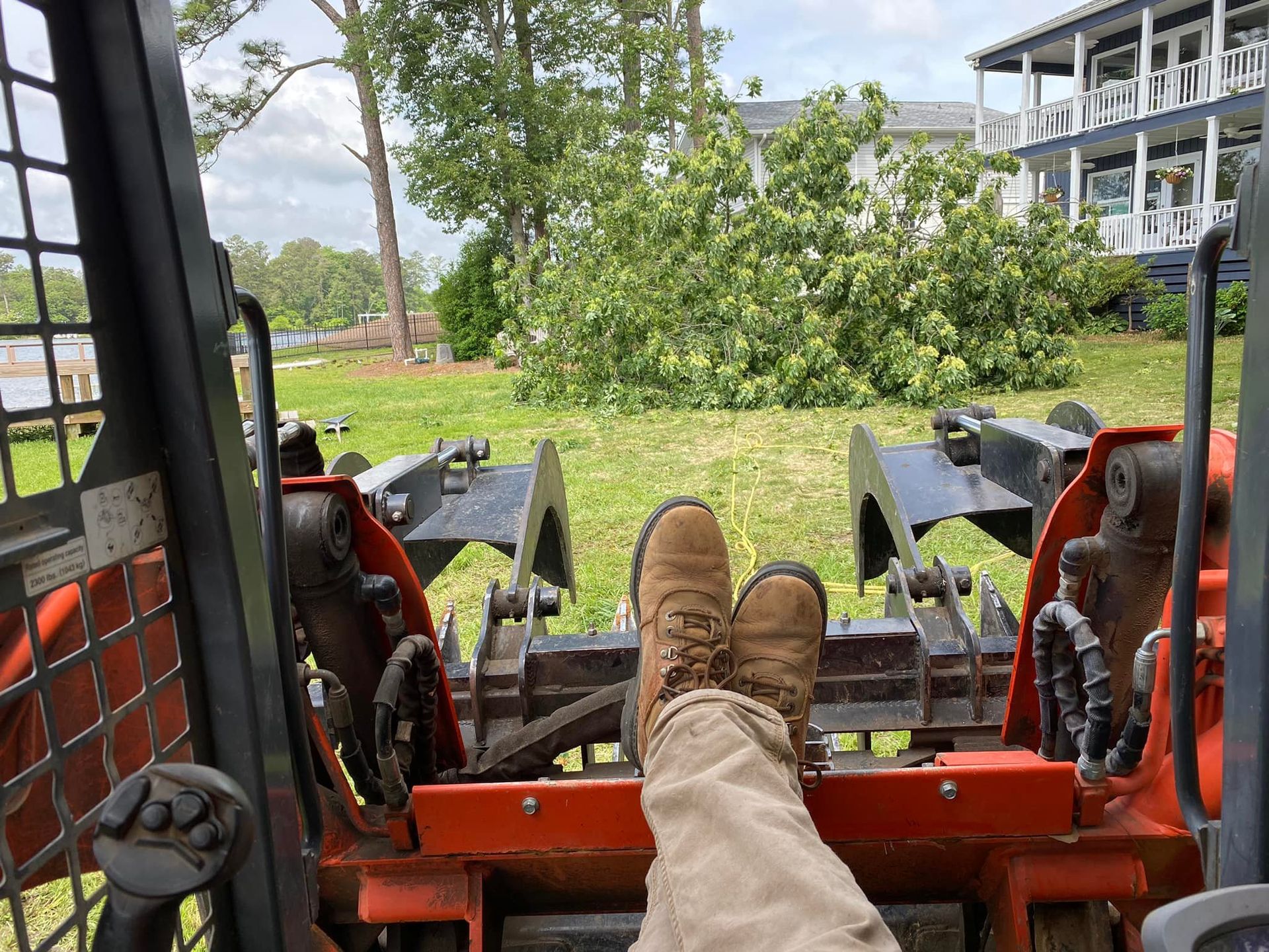 Worker with boots up in a skid steer, looking at fallen tree debris near a house.