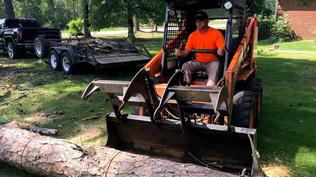 Man operating a skid steer with log grapple; preparing to move a log.