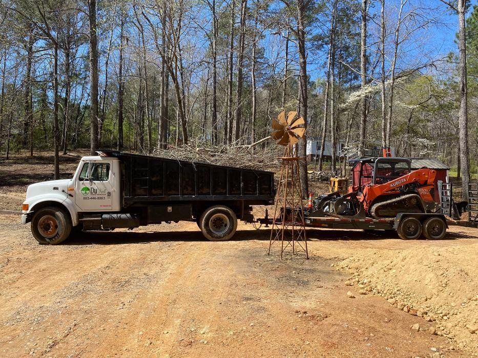 A white dump truck pulling a trailer with an orange skid steer, on a dirt road, trees in background.