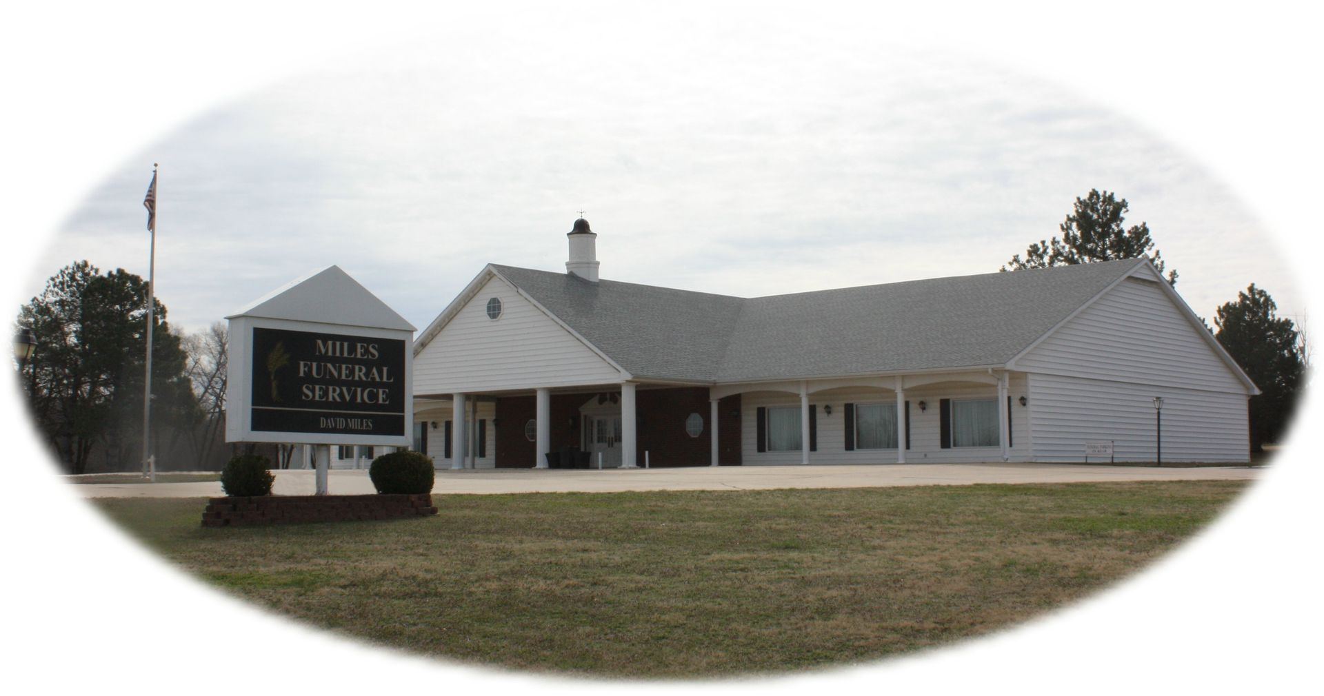 A white funeral home building with a grey roof and a sign in the front lawn against a cloudy sky.