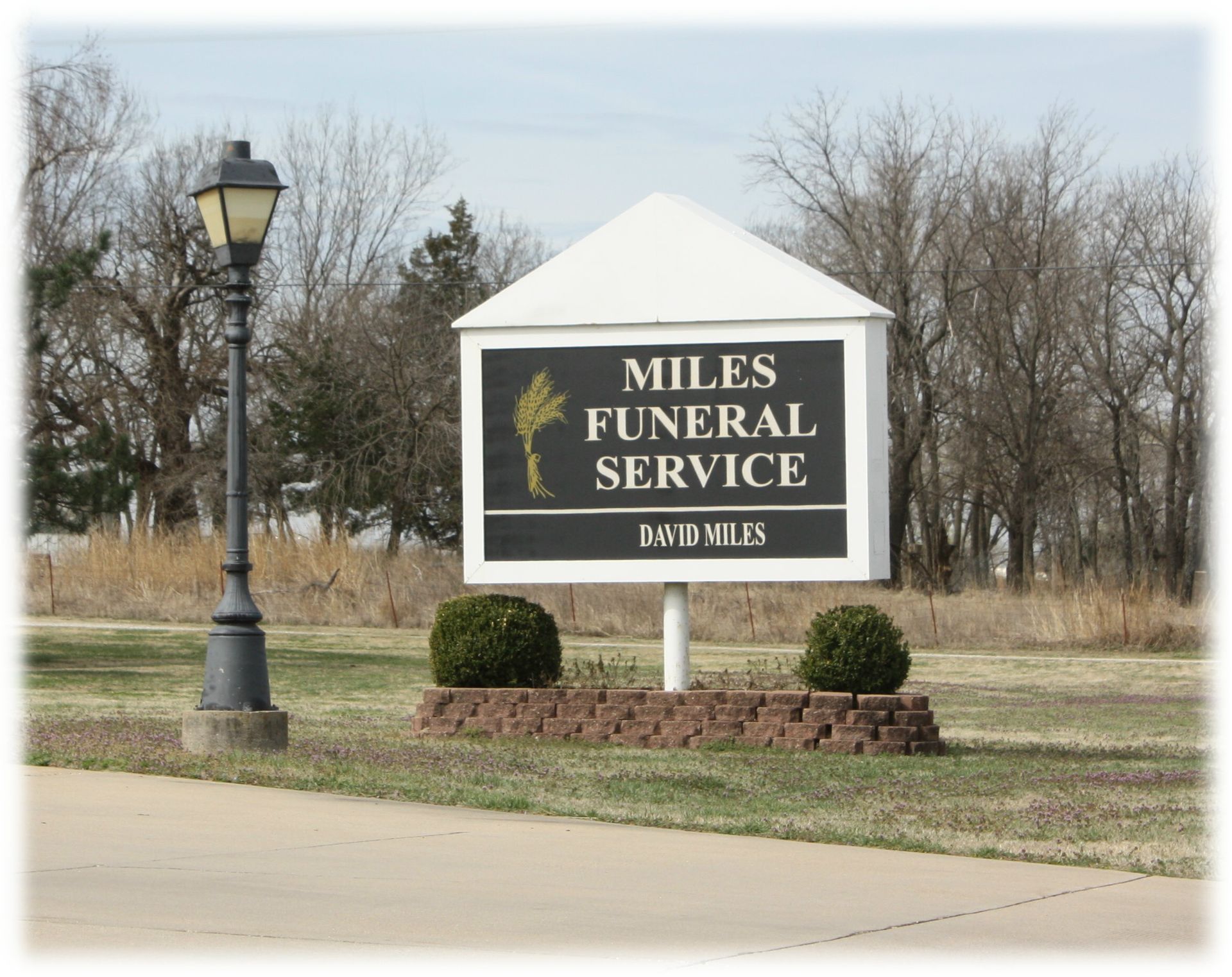 A white, gabled sign for Miles Funeral Service with black lettering and a gold wheat icon, next to a black lamp post.