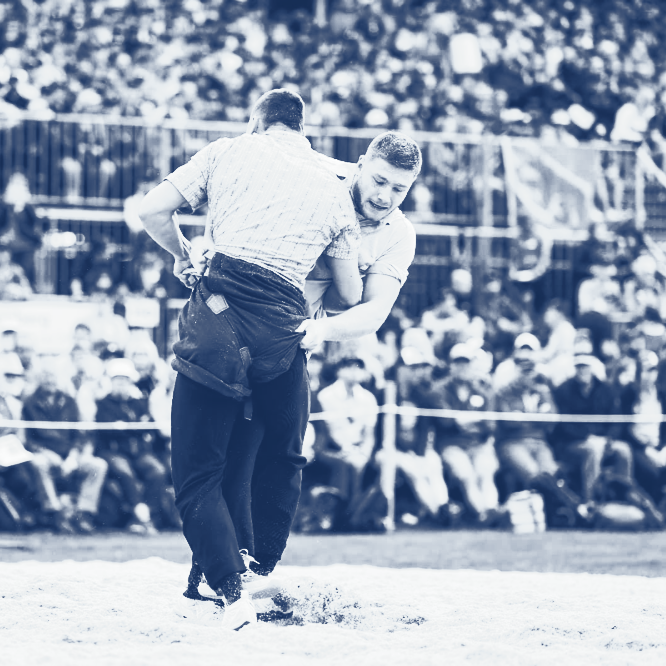 Zwei Männer Schwingen im Sägemehl, umringt von Zuschauern. 