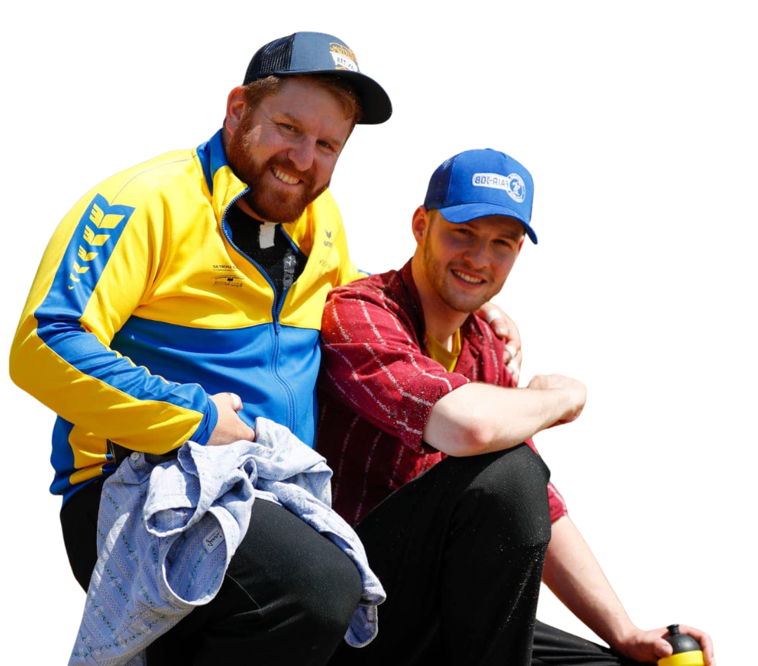 Zwei lächelnde Männer, einer in einer blau-gelben Jacke, der andere in einem rot-karierten Hemd, im Freien, sonniger Tag.