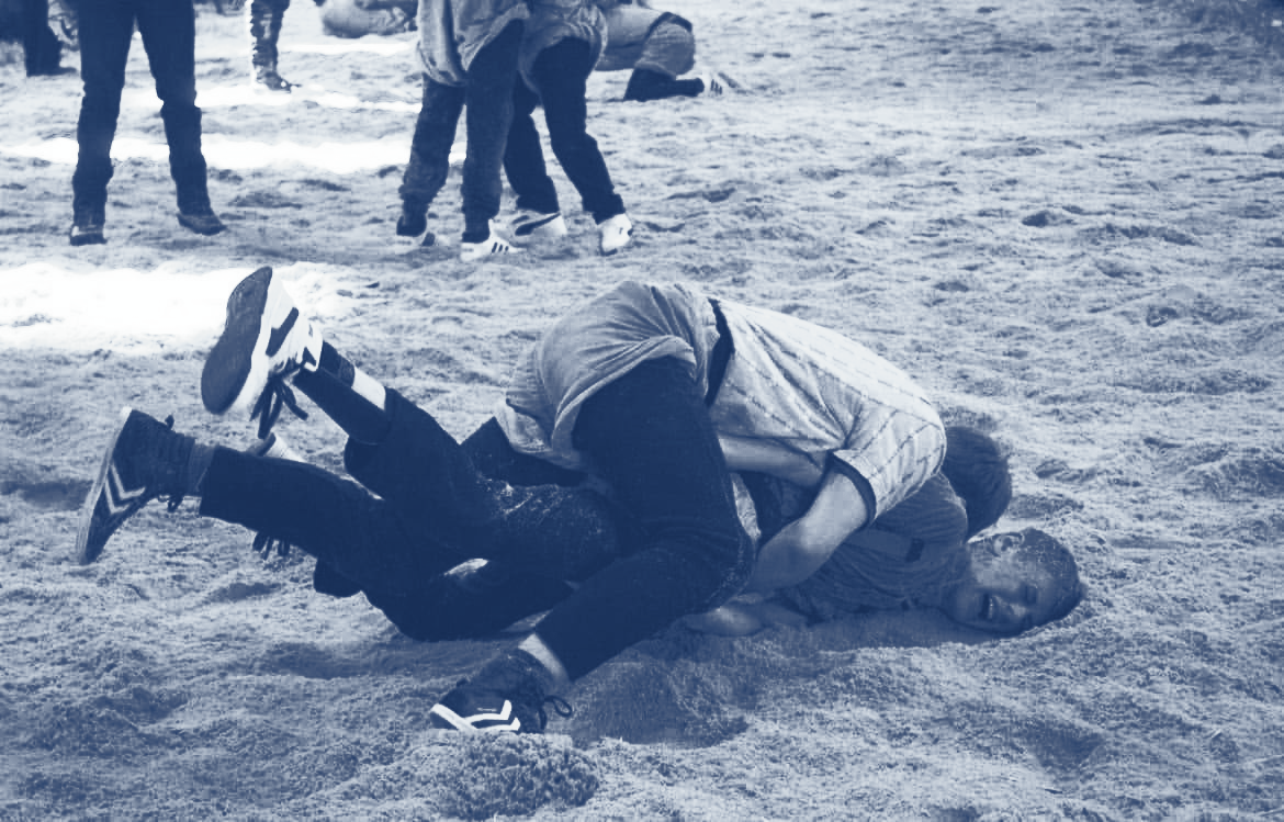 Zwei Personen ringen im Sägemehl, weitere Personen im Hintergrund.