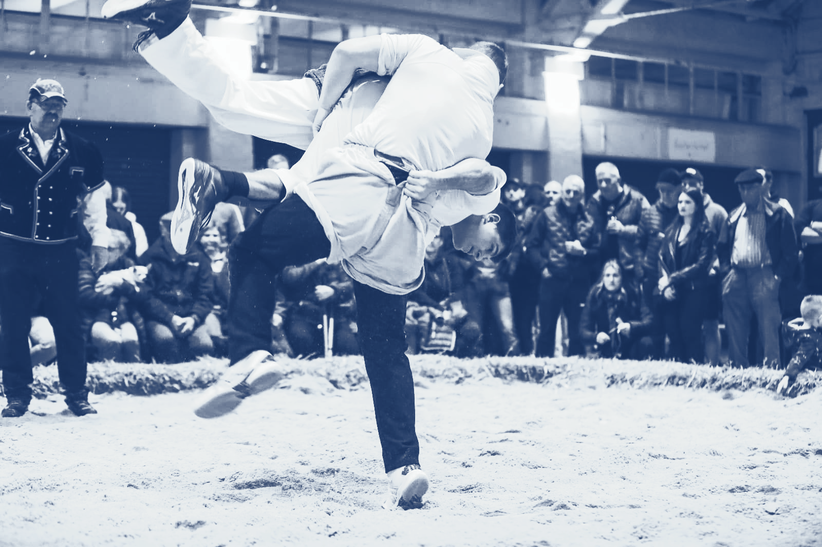 Zwei Schwinger im Sägemehl, einer hebt den anderen mit einem Wurf hoch. Zuschauer beobachten das Geschehen in der Nähe.