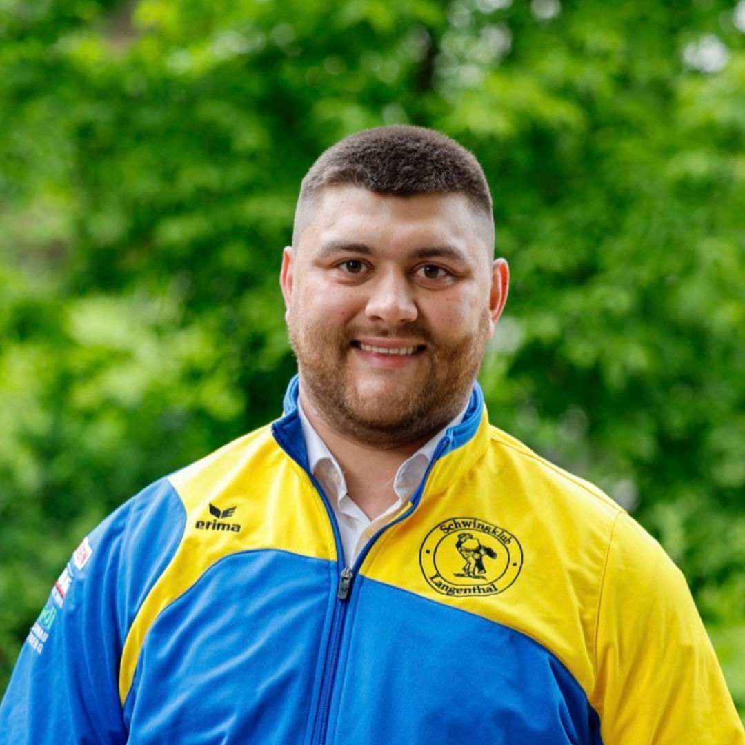 Ein Mann in einer blau-gelben Jacke lächelt vor einem verschwommenen grünen Hintergrund.