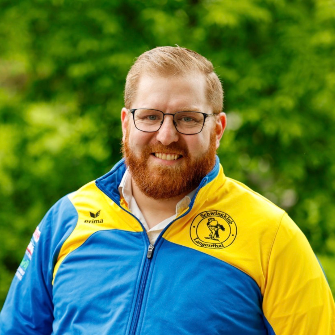 Mann mit rotem Bart, Brille und blau-gelber Jacke, lächelt im Freien.