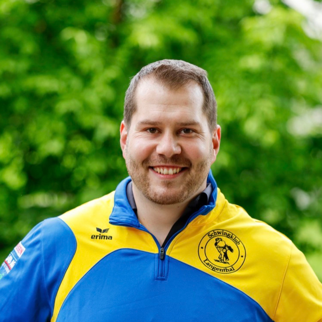Ein lächelnder Mann in einer blau-gelben Sportjacke, im Hintergrund grünes Laub.