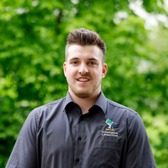 Mann mit braunen Haaren und grauem Hemd lächelt vor grünem Laub.