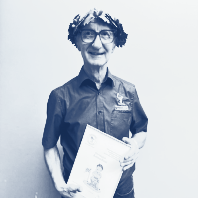 Person mit Lorbeerkranz und Brille hält eine Urkunde und lächelt. Dunkles Hemd, heller Hintergrund.