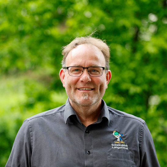Ein Mann mit Brille, grauem Hemd und kleinem Spitzbart lächelt vor grünem Laub.