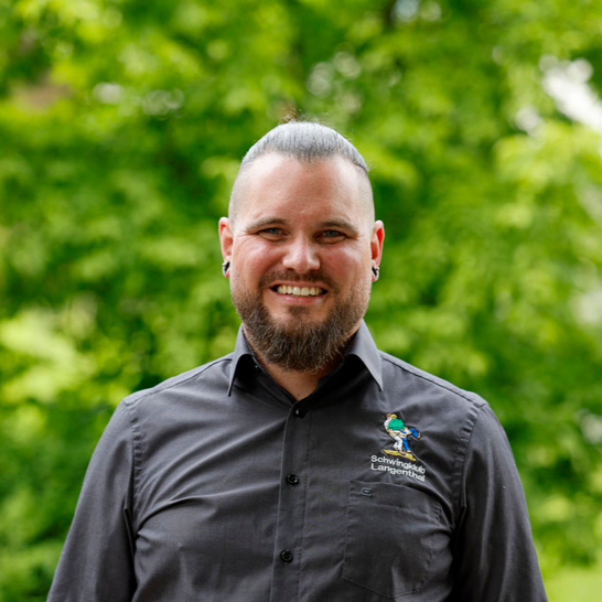 Ein Mann mit Bart lächelt vor grünem Laubwerk; er trägt ein graues Hemd mit Logo.