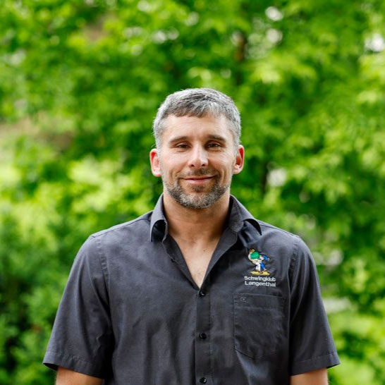 Ein Mann lächelt vor grünem Laubwerk. Er trägt ein dunkelgraues Hemd mit Logo.