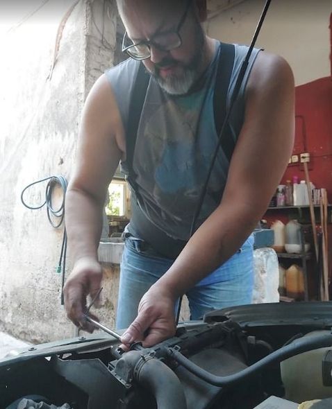 Hombre con camiseta sin mangas trabajando bajo el capó de un coche en un garaje.