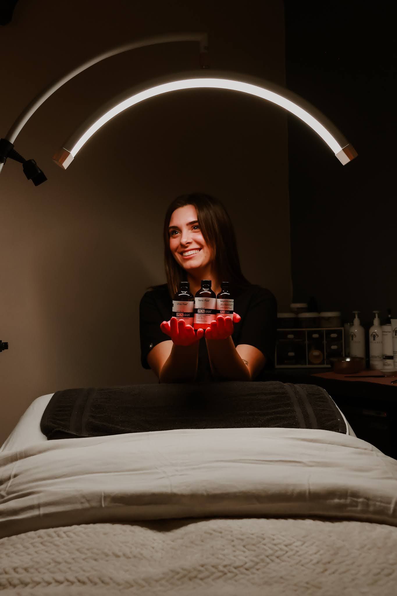 A woman is standing in front of a massage table holding red bottles.