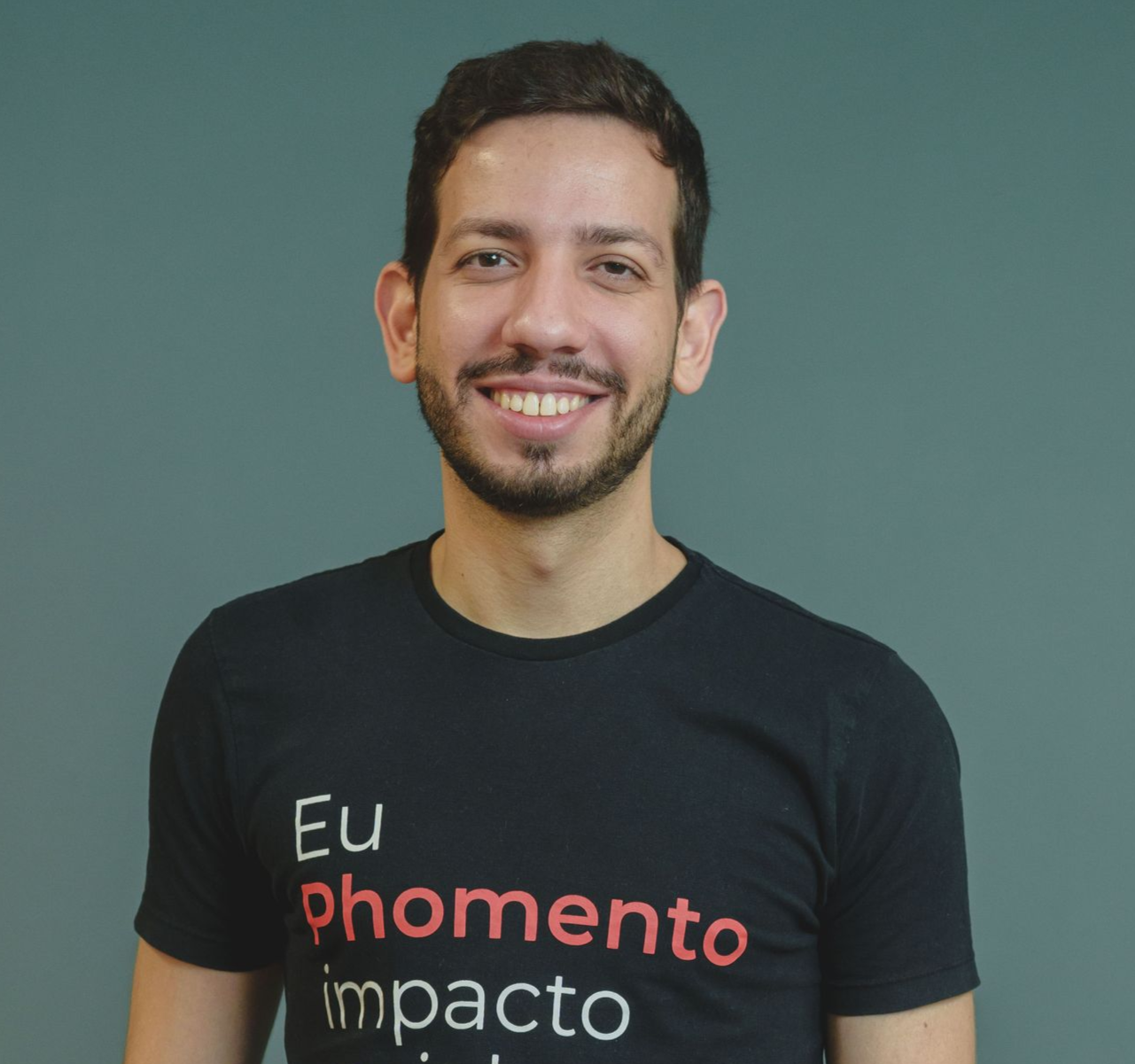 Homem com cabelo e barba escuros sorri para a câmera, vestindo uma camiseta preta.