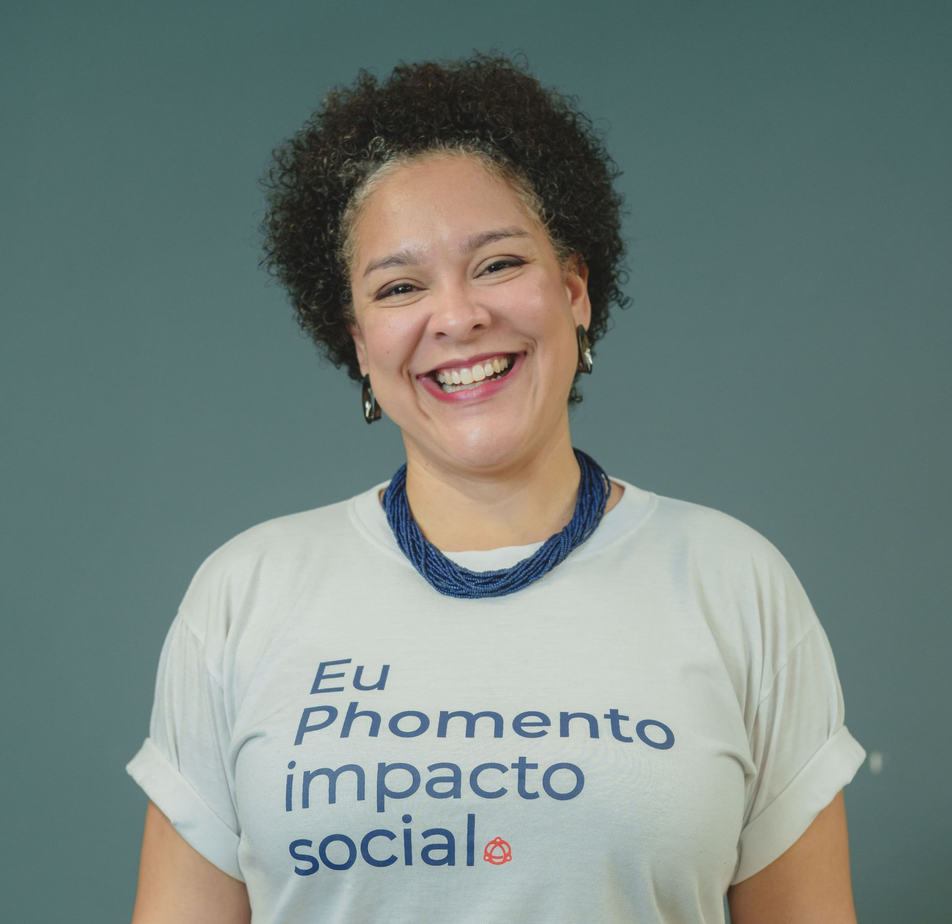 Mulher com cabelos cacheados, sorrindo, vestindo uma camisa branca com texto.