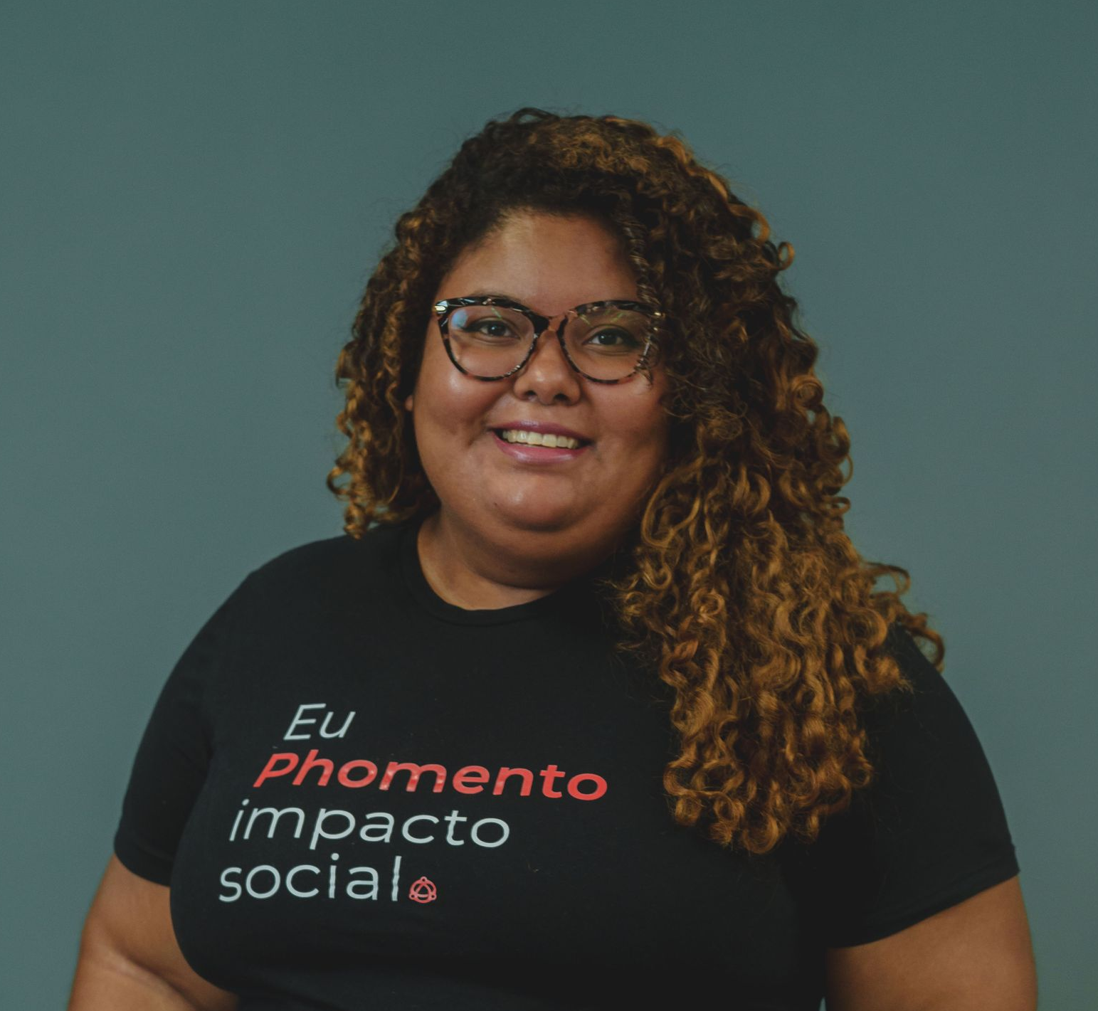 Mulher com cabelos castanhos cacheados sorri, vestindo uma camisa branca com texto, contra um fundo branco.