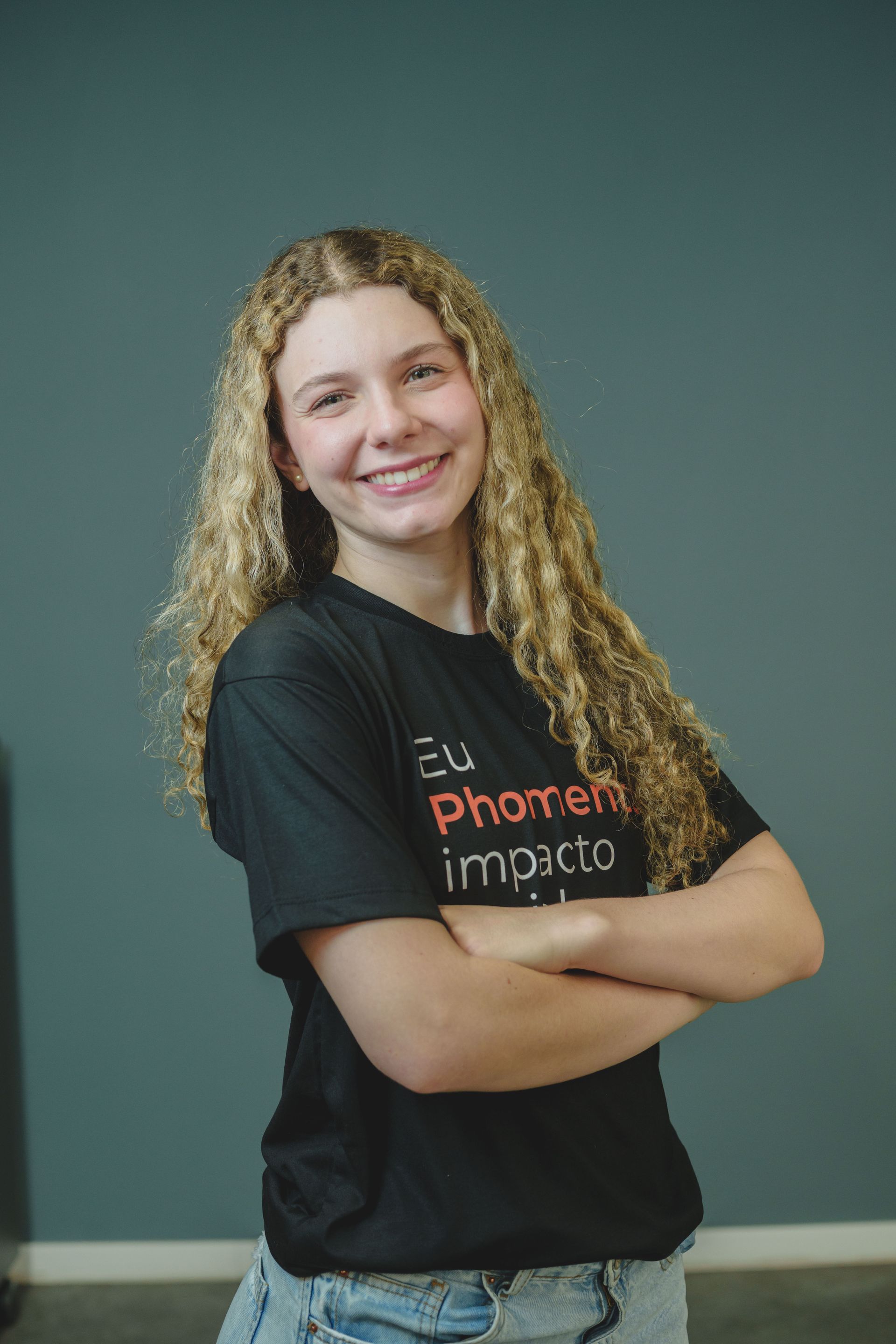 Pessoa sorridente com cabelo loiro encaracolado, vestindo uma camiseta preta com estampa vermelha, em pé contra uma parede verde-azulada escura.