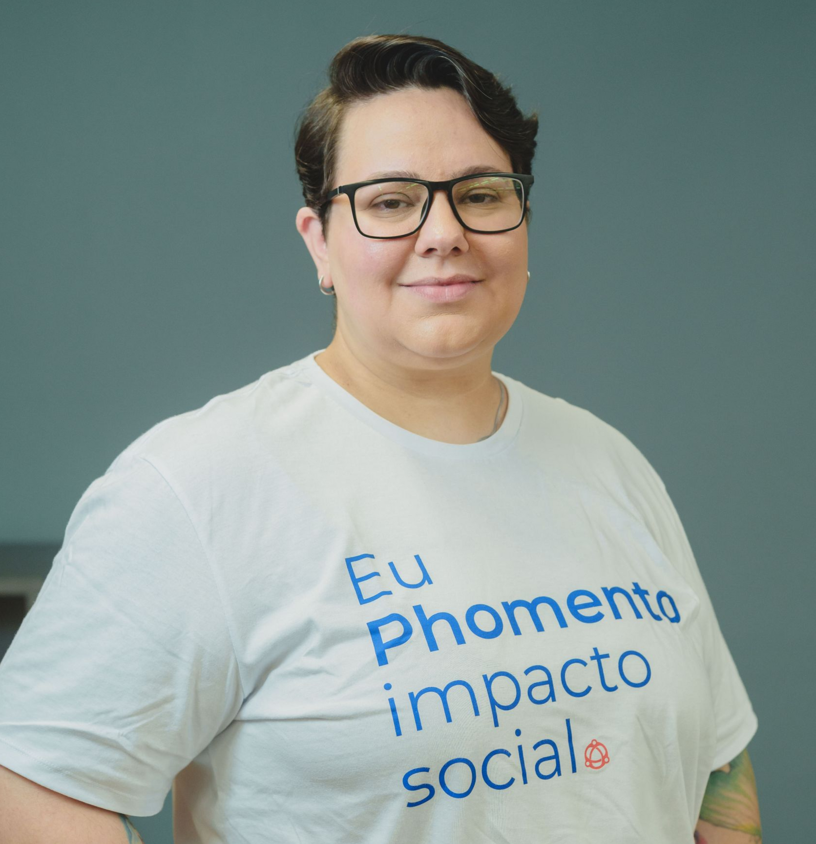 Pessoa sorridente com cabelo escuro e cacheado, óculos e camisa preta sobre fundo branco.