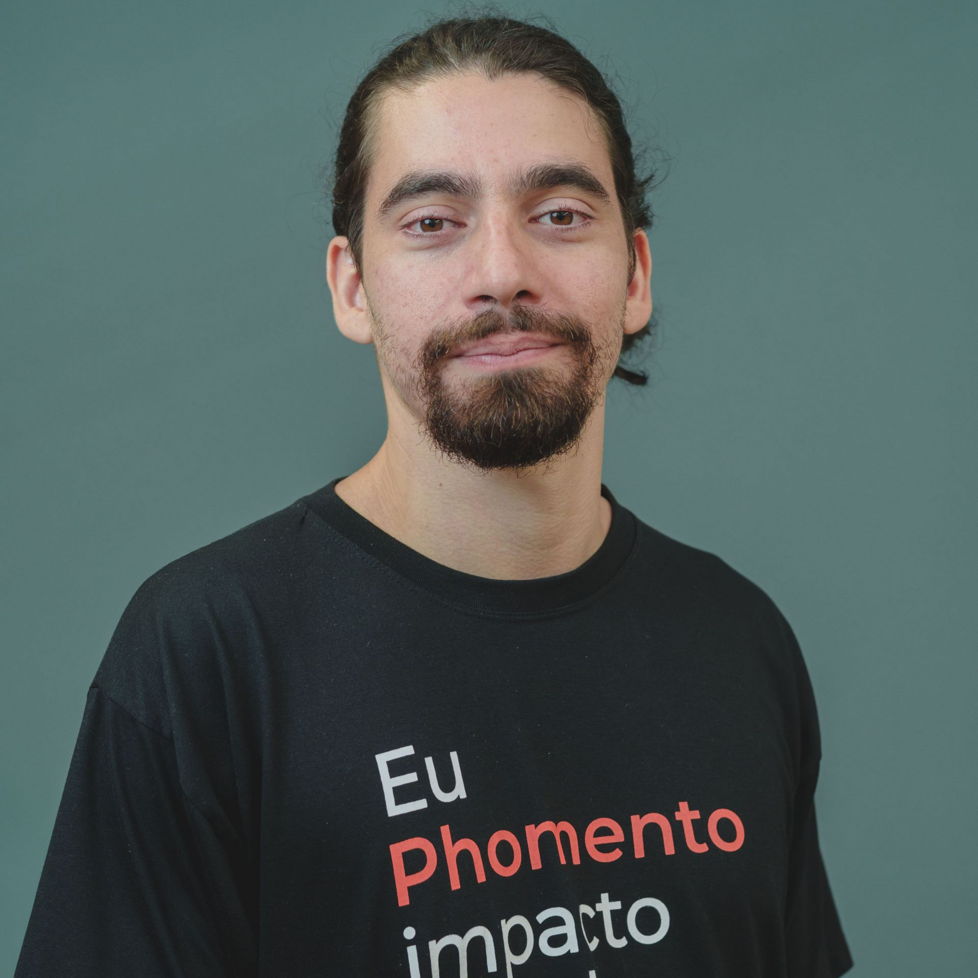 Homem com cabelo e barba escuros sorri, mão no queixo, vestindo camisa preta sobre fundo branco.