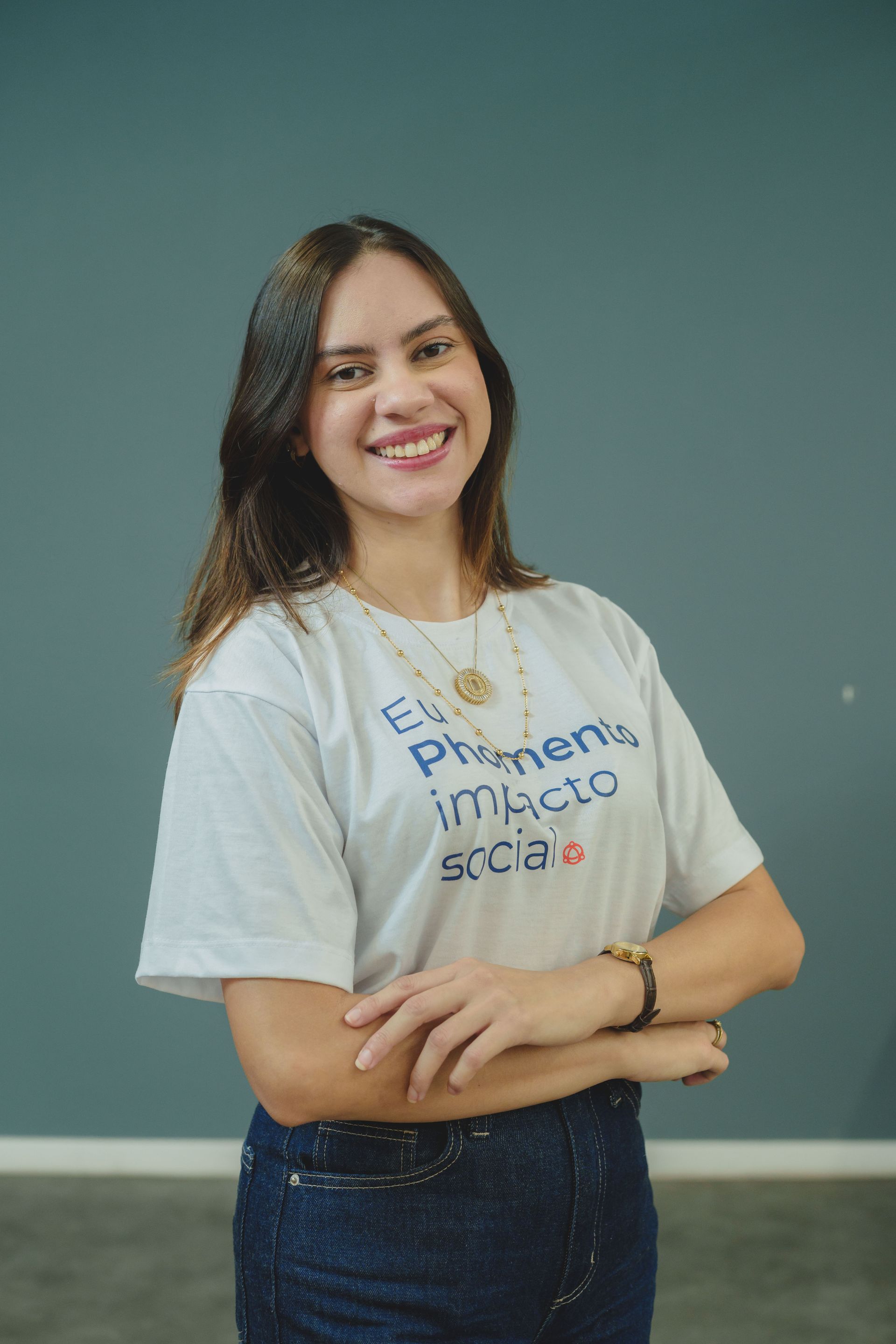 Uma pessoa sorridente está de pé, com os braços cruzados, encostada em uma parede lisa azul-petróleo, vestindo uma camiseta branca e calça jeans escura.