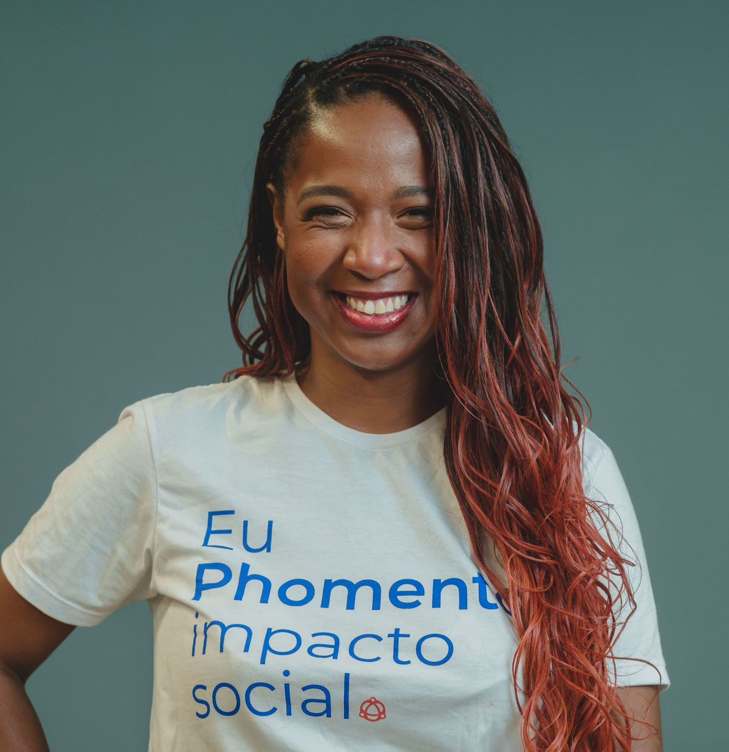 Uma pessoa sorridente, vestindo um suéter branco e com tranças, com óculos escuros pendurados na gola, contra uma parede bege.