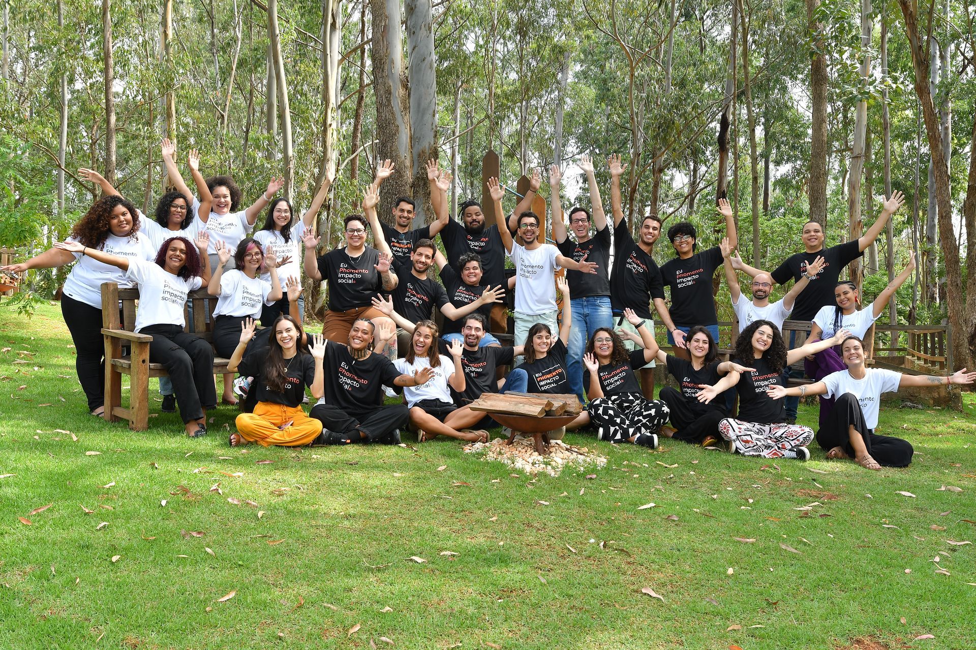 Um grande grupo de pessoas sorrindo e acenando enquanto posam para uma foto em um gramado em uma área arborizada.