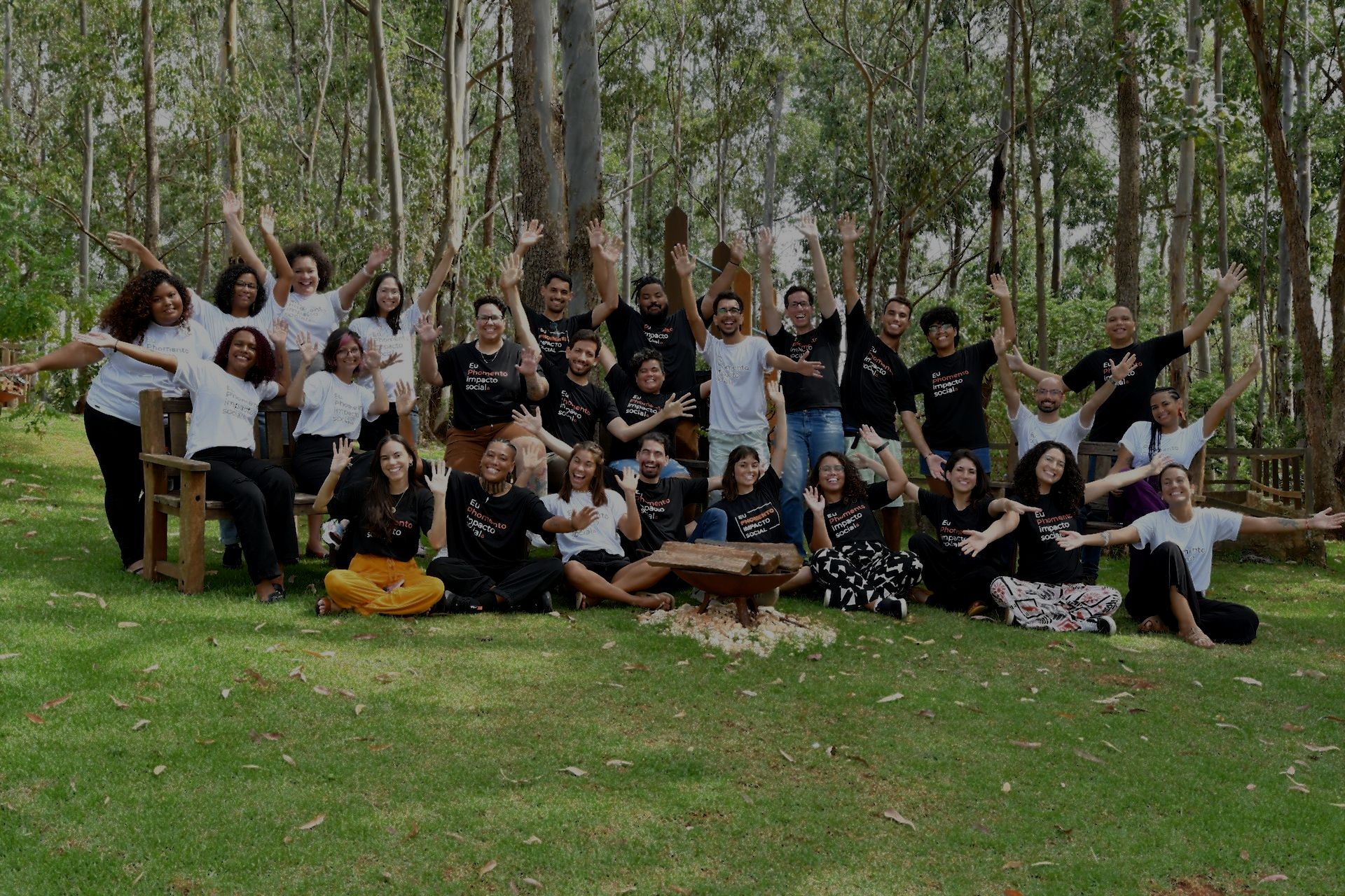 Um grande grupo de pessoas sorrindo e acenando enquanto posam para uma foto em um gramado em uma área arborizada.