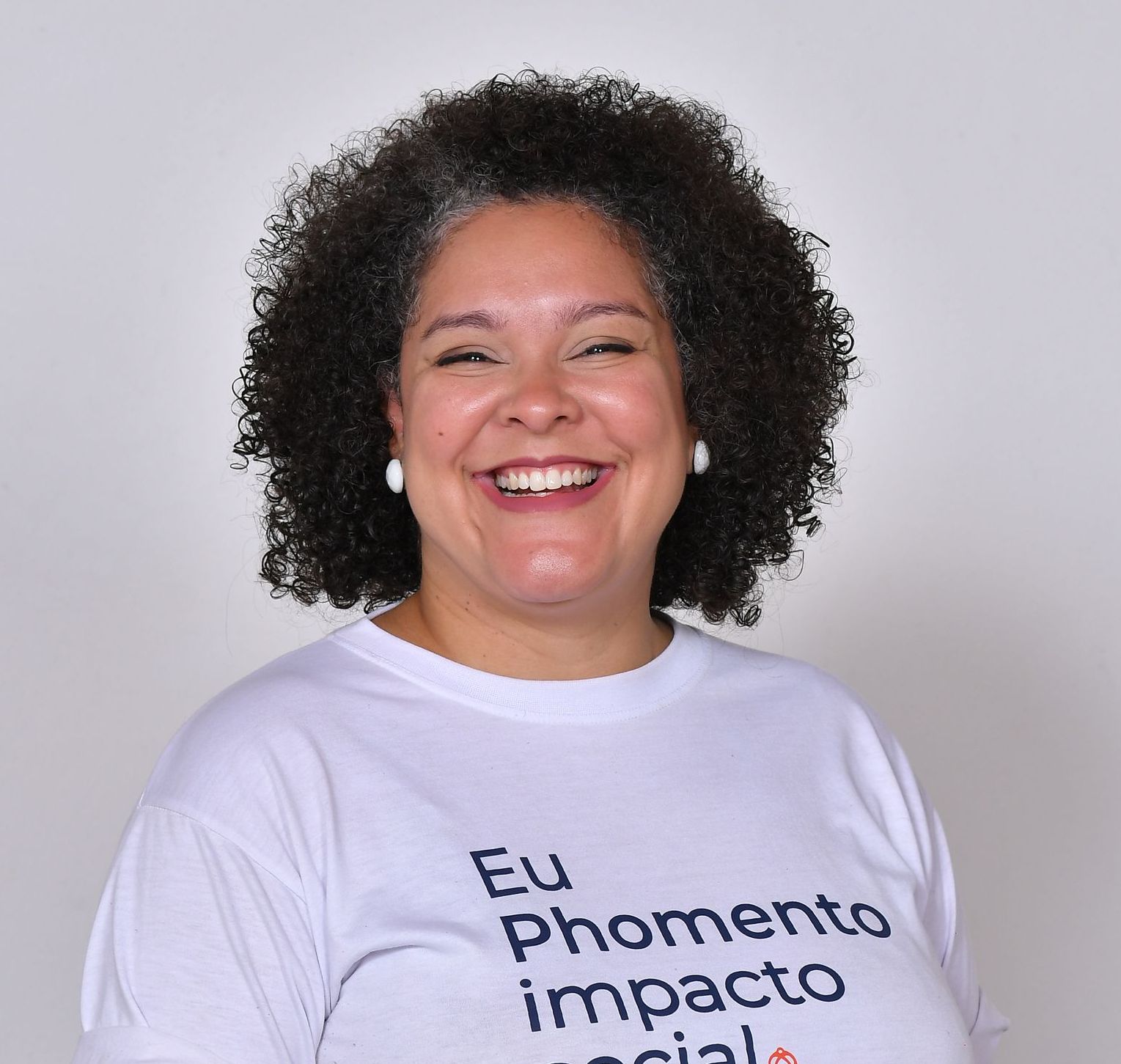 Mulher com cabelos cacheados, sorrindo, vestindo uma camisa branca com texto.
