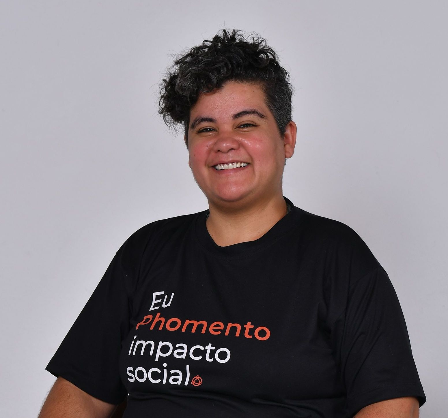 Pessoa sorridente com cabelo curto e cacheado, vestindo uma camiseta preta com os dizeres 