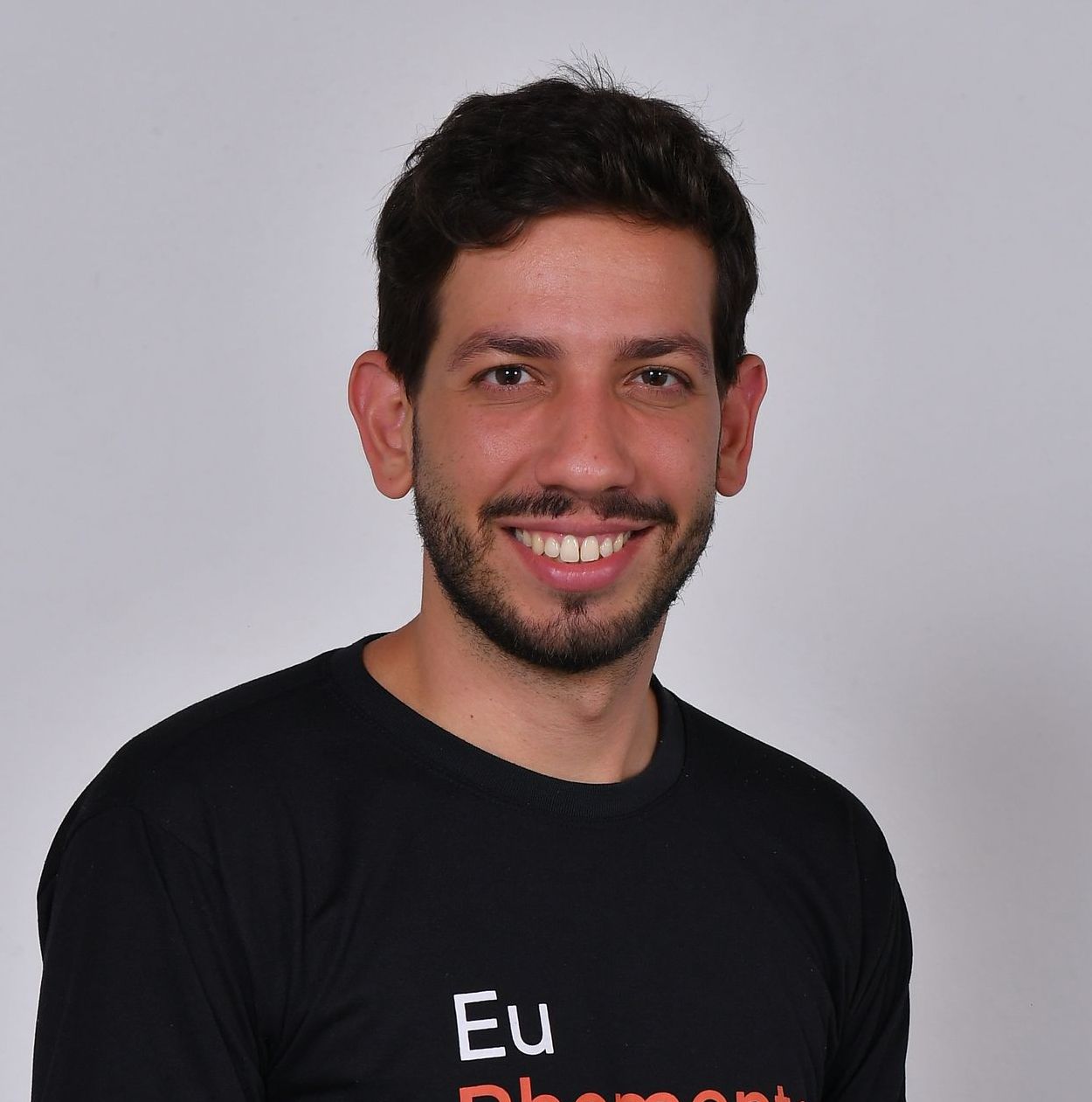 Homem com cabelo e barba escuros sorri para a câmera, vestindo uma camiseta preta.