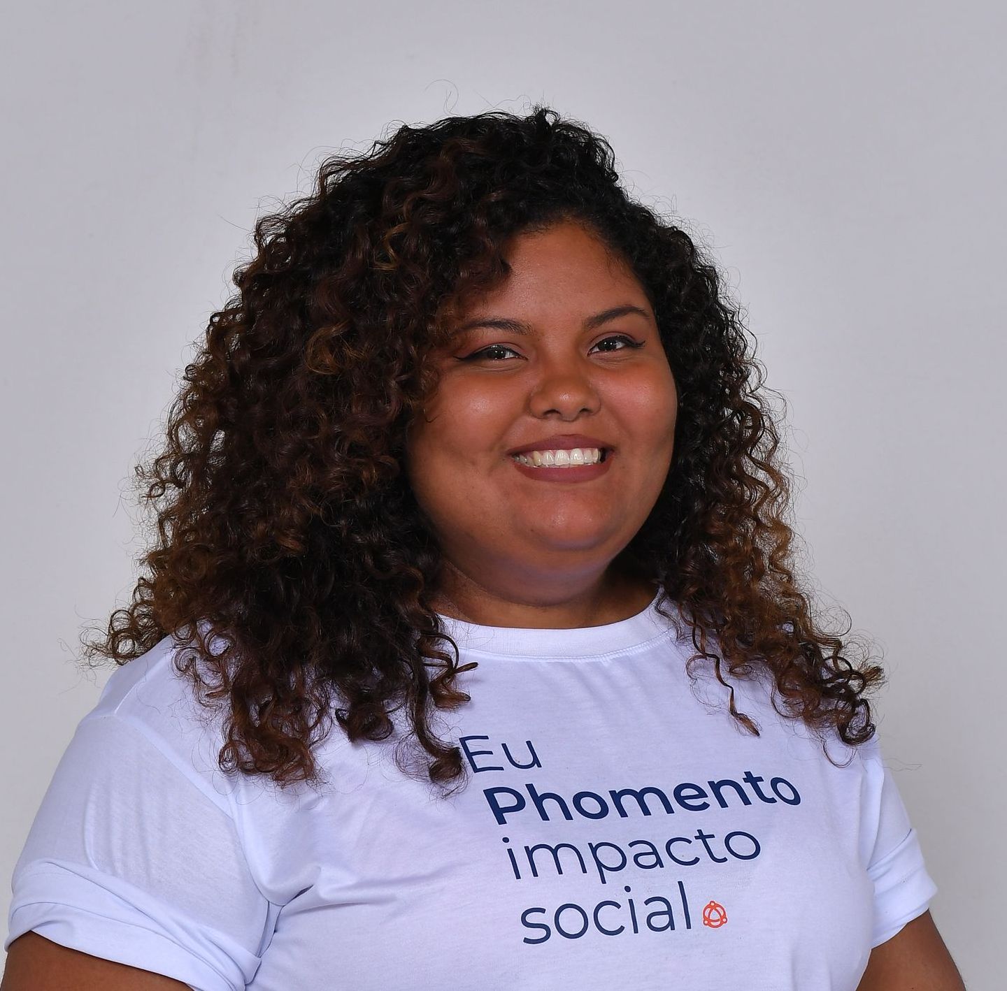 Mulher com cabelos castanhos cacheados sorri, vestindo uma camisa branca com texto, contra um fundo branco.