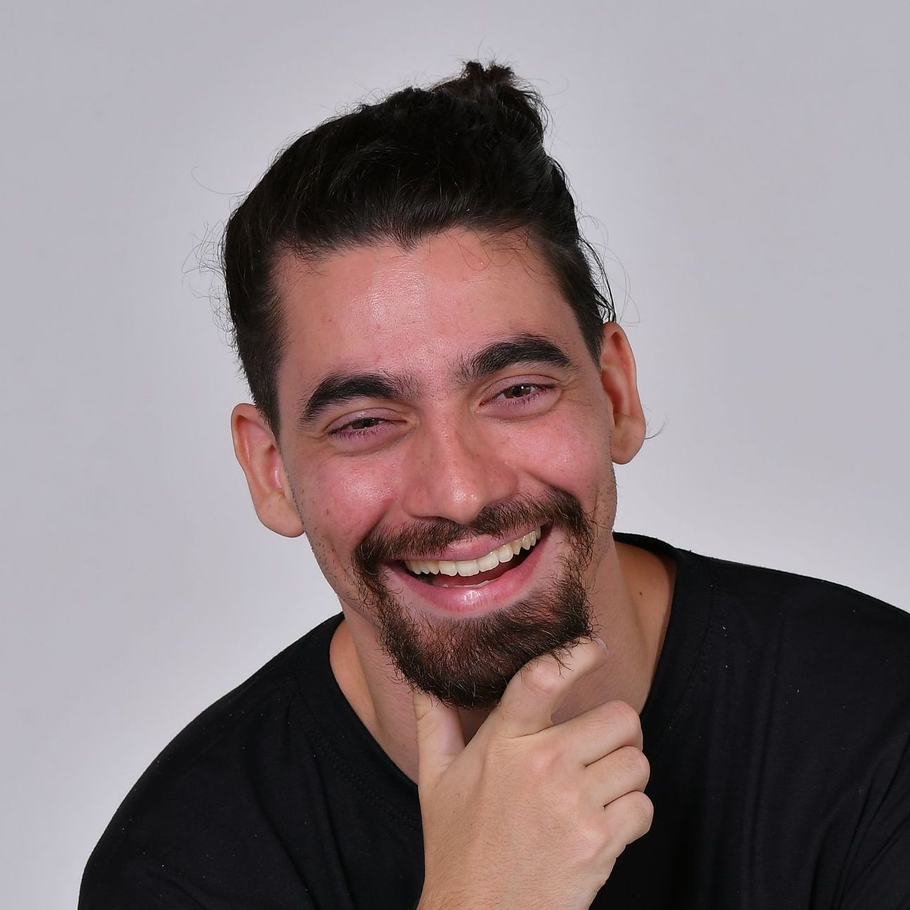 Homem com cabelo e barba escuros sorri, mão no queixo, vestindo camisa preta sobre fundo branco.