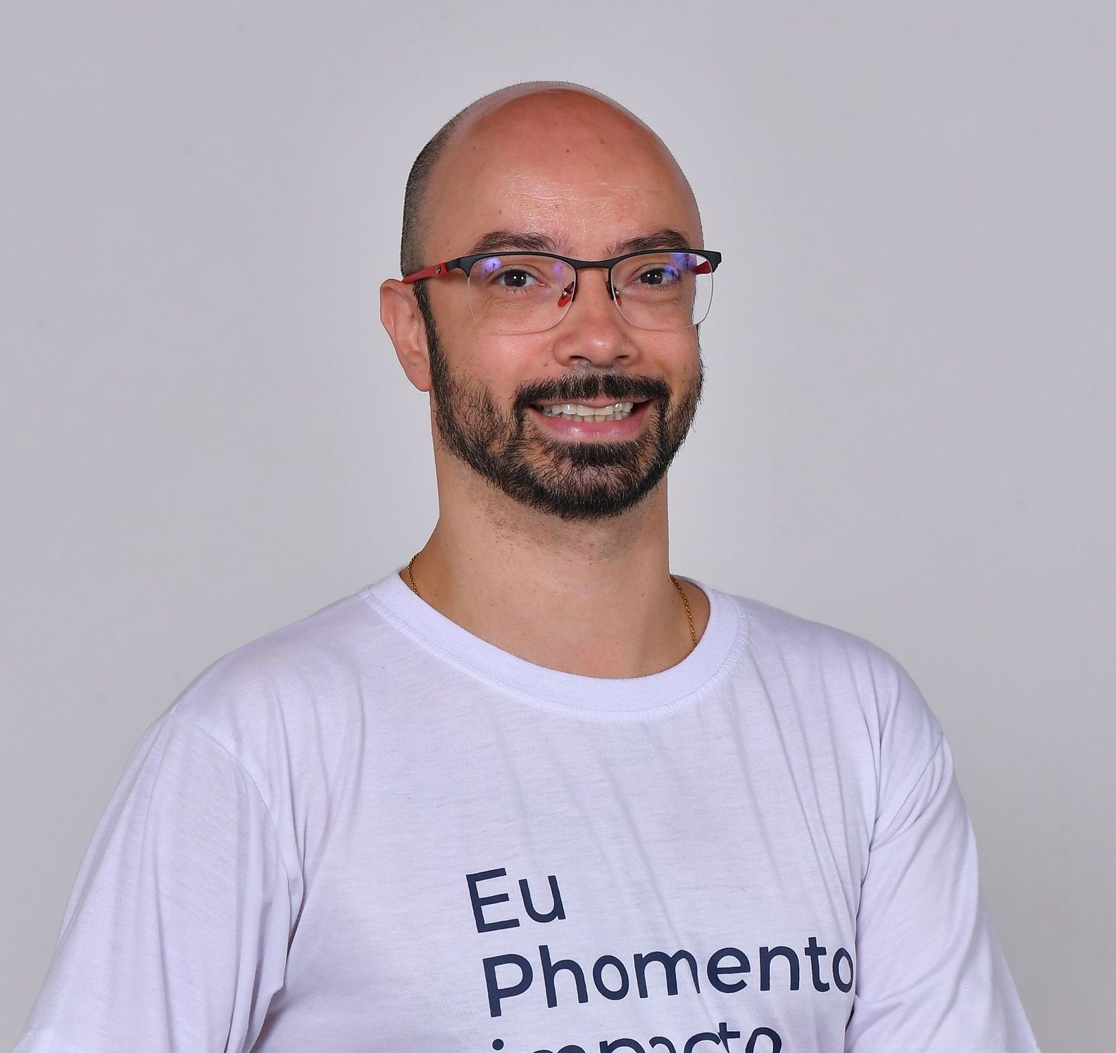Homem de óculos e barba, vestindo uma camiseta branca, sorrindo sobre um fundo cinza claro.
