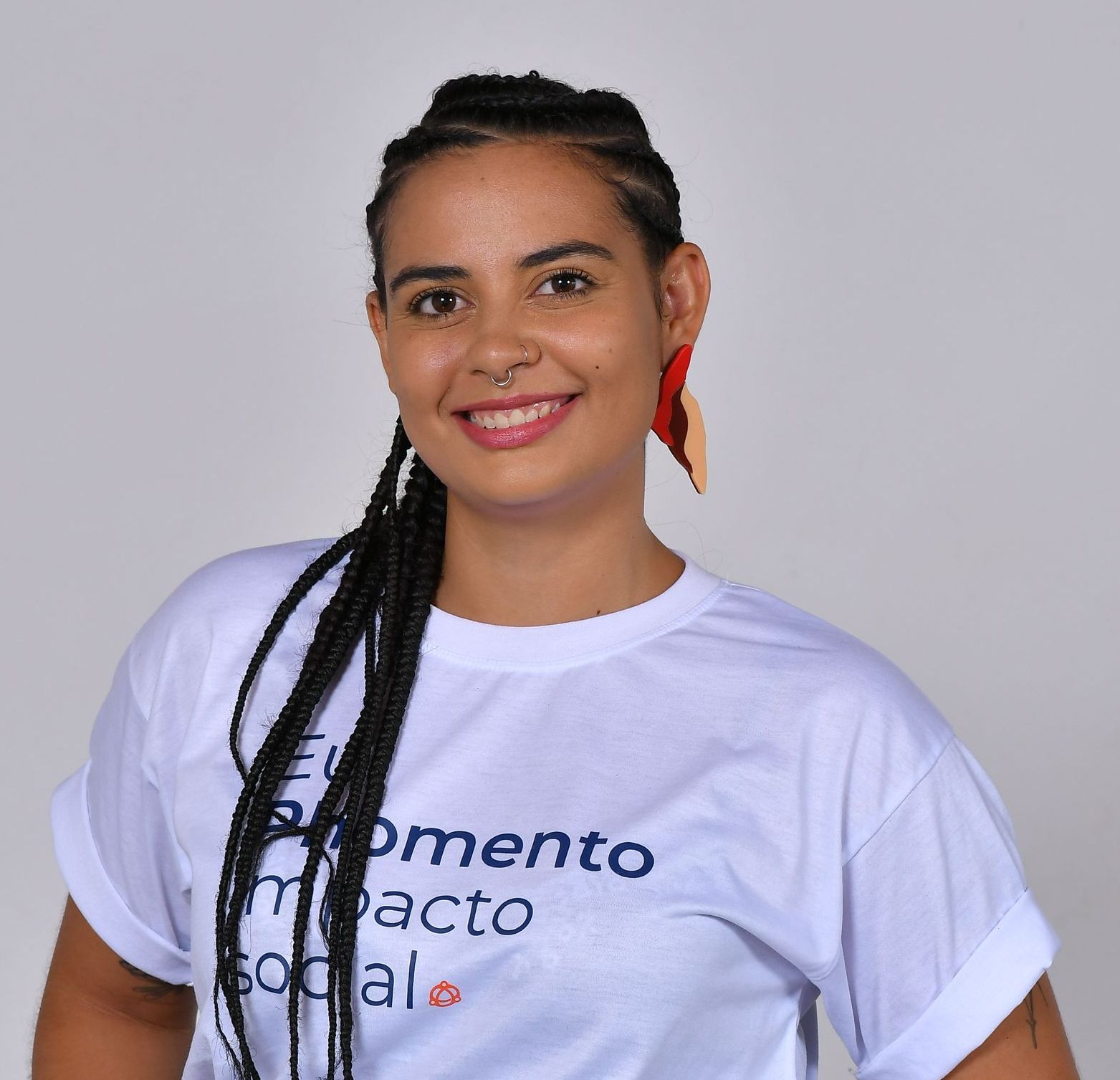 Mulher com tranças escuras, vestindo uma camiseta branca, brincos grandes e um piercing no nariz, sorrindo.