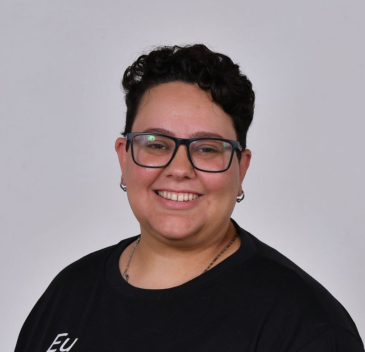 Pessoa sorridente com cabelo escuro e cacheado, óculos e camisa preta sobre fundo branco.